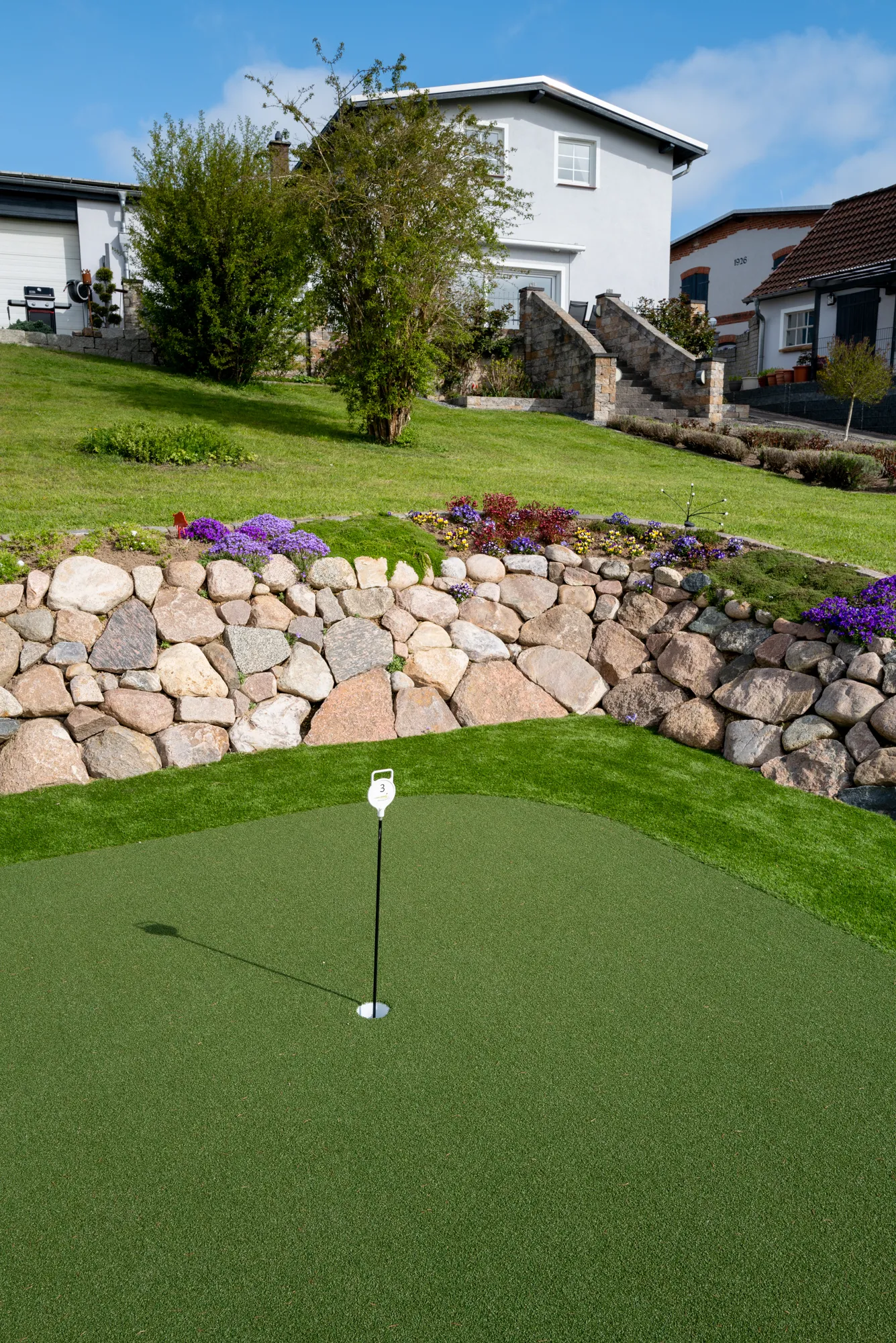 Angelegtes Kunstrasen-Green mit Fahne und Natursteinmauer in einem Hanggarten